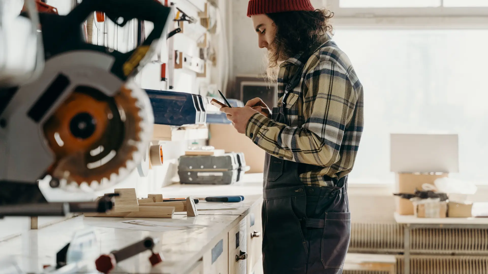 A craftsperson checking a phone in a workshop, with a workbench and tools behind